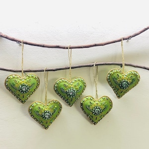 Op de afbeelding: Vijf groene hartvormige ornamenten met gouden randen en kralenaccenten hangen aan een bruine tak. Elk hart heeft een centraal bloemmotief en wordt opgehangen door een gouden koord. Dit zijn feestelijke decoraties.