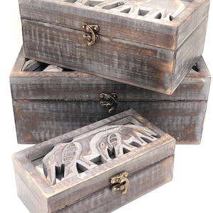 May include: Set of three wooden boxes with elephant carvings. The boxes are gray and have brass clasps.