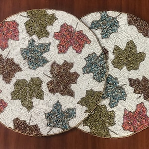Puede incluir: Dos manteles individuales redondos de cuentas blancas con un diseño de hojas. Las hojas son en tonos de marrón, verde, azul y naranja.