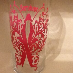 May include: A clear glass with a pink butterfly design and the word "Survivor" in pink lettering. The butterfly has intricate details and a pink ribbon in the center.