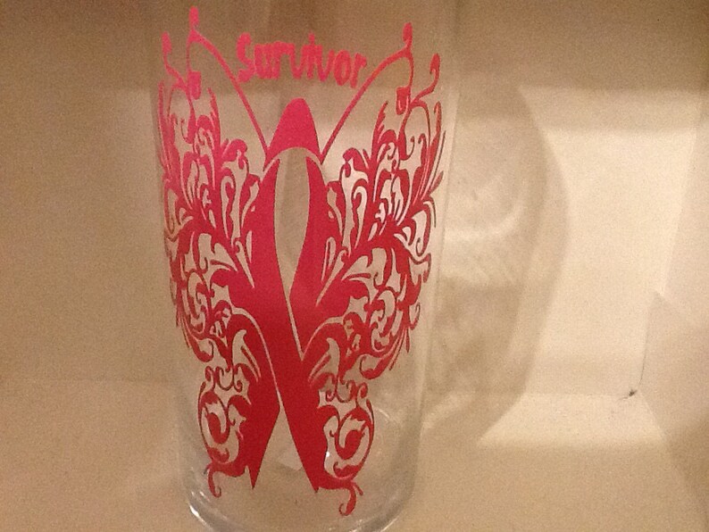 May include: A clear glass with a pink butterfly design and the word "Survivor" in pink lettering. The butterfly has intricate details and a pink ribbon in the center.