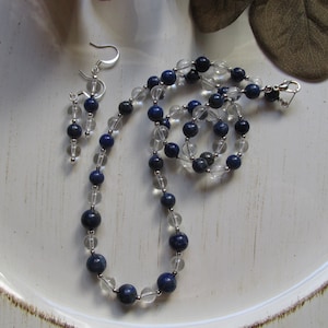 May include: A necklace and earrings set made with clear quartz and lapis lazuli beads. The necklace is silver and has a lobster clasp closure. The earrings are silver and have a hook closure.