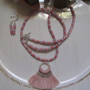 May include: A pink and lavender beaded necklace with a pink tassel pendant and matching earrings. The necklace is made of small, square beads and has a silver clasp.