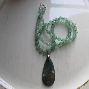 May include: A green jade stone necklace with a teardrop-shaped pendant. The pendant has a mottled green, brown, and white pattern.