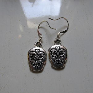 Puede incluir: Pendientes de plata con diseño de calavera de azúcar. La calavera tiene detalles negros y una forma de corazón en la frente.