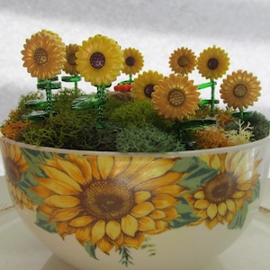 May include: A decorative bowl featuring a sunflower design. The bowl is filled with green moss and artificial yellow sunflowers. The sunflowers have a detailed design and are arranged to create a miniature garden scene.