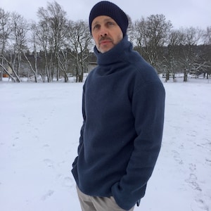 May include: A man wearing a dark blue turtleneck sweater and beige pants stands in a snowy field. He is wearing a dark blue knit beanie.