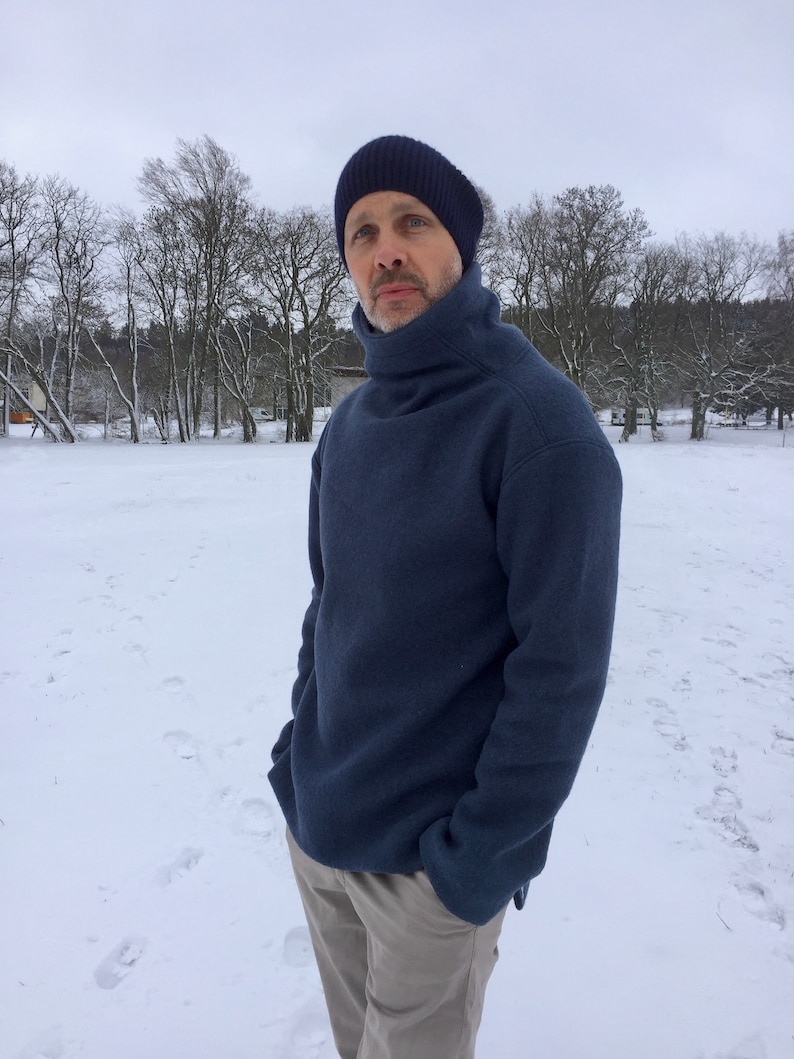 May include: A man wearing a dark blue turtleneck sweater and beige pants stands in a snowy field. He is wearing a dark blue knit beanie.