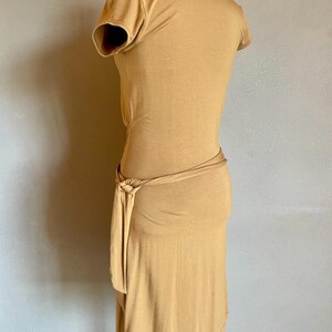 K&ouml;nnte beinhalten: Senffarbenes Kleid mit kurzen &Auml;rmeln und einem Wickel-Taillenband. Das Kleid wird auf einer Schaufensterpuppe pr&auml;sentiert, die sein figurbetontes Design zeigt. Der Stoff scheint ein weiches, jerseyartiges Material zu sein.