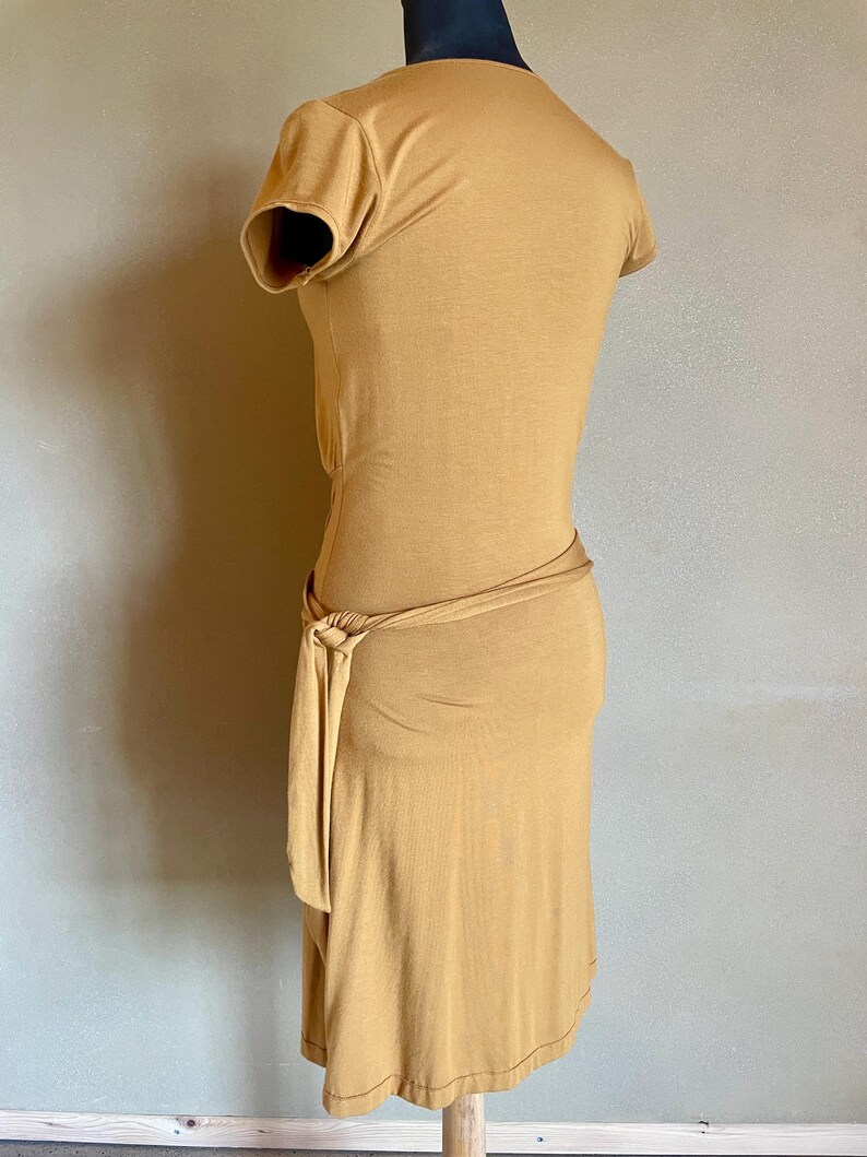 K&ouml;nnte beinhalten: Senffarbenes Kleid mit kurzen &Auml;rmeln und einem Wickel-Taillenband. Das Kleid wird auf einer Schaufensterpuppe pr&auml;sentiert, die sein figurbetontes Design zeigt. Der Stoff scheint ein weiches, jerseyartiges Material zu sein.