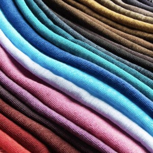 K&ouml;nnte beinhalten: Nahaufnahme einer Sammlung farbenfroher Stoffmuster, die diagonal angeordnet sind. Die Muster zeigen eine gro&szlig;e Auswahl an Farben, darunter Rot, Rosa, Blau, Gr&uuml;n und Braun. Der Stoff scheint ein weiches Strickmaterial zu sein.