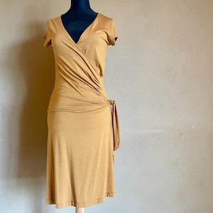 K&ouml;nnte beinhalten: Senffarbenes Wickelkleid mit kurzen &Auml;rmeln und V-Ausschnitt. Das Kleid hat ein drapiertes Design &uuml;ber dem Oberteil und ein Bindedetail an der Taille. Das Kleid wird auf einer schwarzen Schaufensterpuppe pr&auml;sentiert.