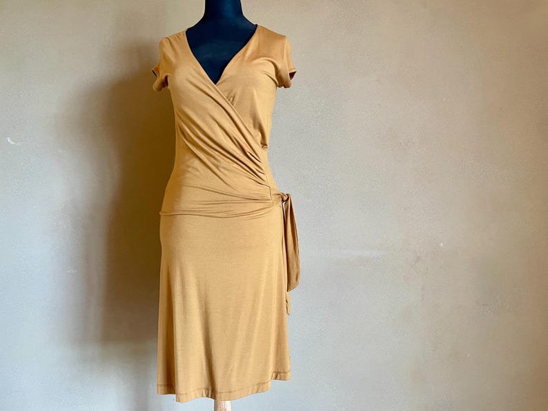 K&ouml;nnte beinhalten: Senffarbenes Wickelkleid mit kurzen &Auml;rmeln und V-Ausschnitt. Das Kleid hat ein drapiertes Design &uuml;ber dem Oberteil und ein Bindedetail an der Taille. Das Kleid wird auf einer schwarzen Schaufensterpuppe pr&auml;sentiert.