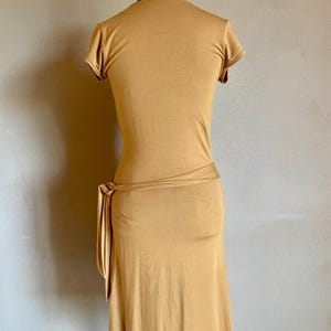 K&ouml;nnte beinhalten: Senffarbenes Kleid mit kurzen &Auml;rmeln und einem Wickelband in der Taille. Das Kleid wird auf einer Schaufensterpuppe pr&auml;sentiert, die seine figurbetonte Silhouette und den knielangen Saum zeigt. Der Stoff scheint ein weiches, jerseyartiges Material zu sein.