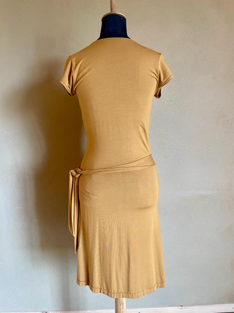 K&ouml;nnte beinhalten: Senffarbenes Kleid mit kurzen &Auml;rmeln und einem Wickelband in der Taille. Das Kleid wird auf einer Schaufensterpuppe pr&auml;sentiert, die seine figurbetonte Silhouette und den knielangen Saum zeigt. Der Stoff scheint ein weiches, jerseyartiges Material zu sein.