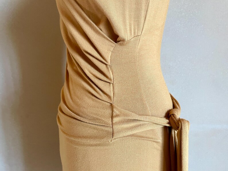 K&ouml;nnte beinhalten: Ein hellbraunes Kleid mit drapiertem Oberteil und einem geknoteten Taillendetail. Das Kleid hat eine figurbetonte Silhouette und scheint aus einem weichen, dehnbaren Stoff gefertigt zu sein. Das Design ist schlicht und elegant.