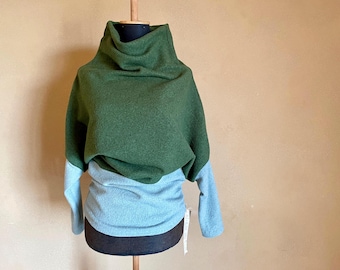 Puede incluir: Un suéter de dos tonos en tonos verdes y azules, exhibido en un maniquí. La parte superior es de un verde oscuro, que pasa a un azul más claro en las mangas y la parte inferior del cuerpo. El suéter tiene un cuello alto y drapeado.