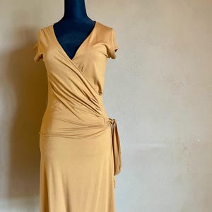 K&ouml;nnte beinhalten: Ein senffarbenes Wickelkleid mit kurzen &Auml;rmeln und V-Ausschnitt. Das Kleid hat ein gerafftes Detail an der Seite und einen Bindverschluss. Das Kleid wird auf einer schwarzen Schaufensterpuppe vor neutralem Hintergrund pr&auml;sentiert.