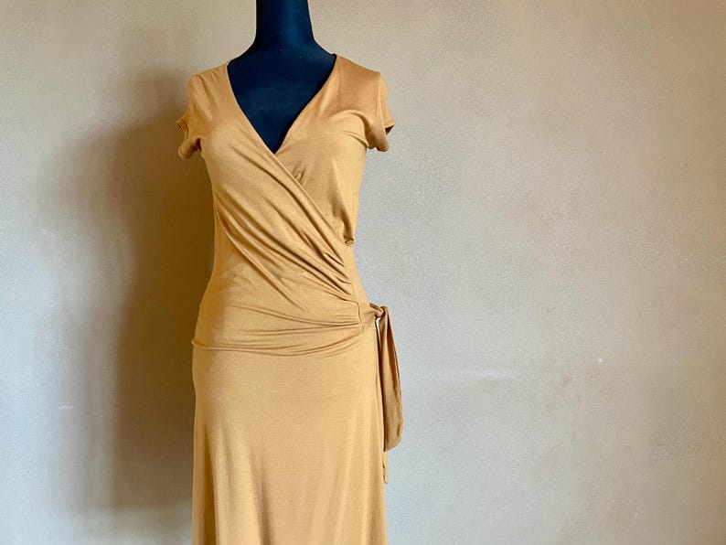 K&ouml;nnte beinhalten: Ein senffarbenes Wickelkleid mit kurzen &Auml;rmeln und V-Ausschnitt. Das Kleid hat ein gerafftes Detail an der Seite und einen Bindverschluss. Das Kleid wird auf einer schwarzen Schaufensterpuppe vor neutralem Hintergrund pr&auml;sentiert.