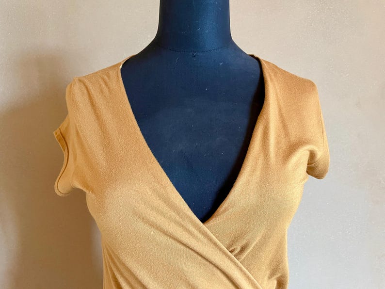 K&ouml;nnte beinhalten: Senffarbenes Wickeloberteil mit kurzen &Auml;rmeln. Das Oberteil hat einen tiefen V-Ausschnitt und wird auf einer schwarzen Schaufensterpuppe pr&auml;sentiert. Der Stoff scheint einen leichten Glanz zu haben.