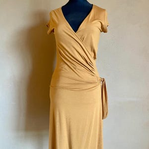 K&ouml;nnte beinhalten: Senffarbenes Wickelkleid mit kurzen &Auml;rmeln und V-Ausschnitt. Das Kleid hat ein drapiertes Design an der Taille und ein Bindedetail an der Seite. Das Kleid wird auf einer schwarzen Schaufensterpuppe pr&auml;sentiert.