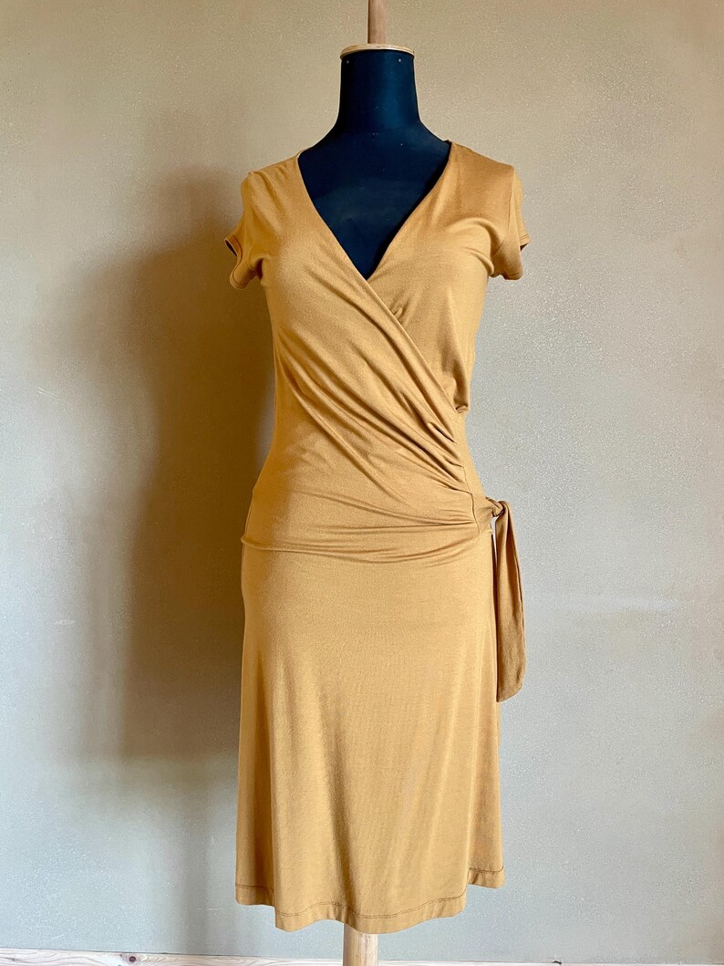 K&ouml;nnte beinhalten: Senffarbenes Wickelkleid mit kurzen &Auml;rmeln und V-Ausschnitt. Das Kleid hat ein drapiertes Design an der Taille und ein Bindedetail an der Seite. Das Kleid wird auf einer schwarzen Schaufensterpuppe pr&auml;sentiert.