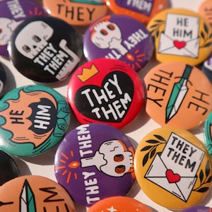 May include: A collection of colourful button badges with various designs and messages. The badges feature illustrations of skulls, hearts, swords, and envelopes. The text on the badges includes "They Them", "He Him", and "They Them".