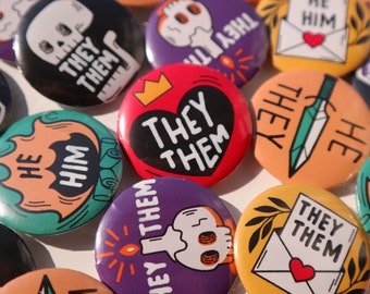 May include: A collection of colorful button pins with various designs and messages. The pins feature illustrations of skulls, hearts, swords, and envelopes. The text on the pins includes "They Them", "He Him", and "They Them".