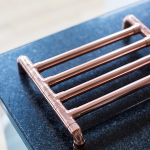 May include: A copper pipe trivet with a rectangular design. The trivet is made of copper pipes joined together to create a sturdy base for hot pots and pans. The copper has a shiny, reflective surface.
