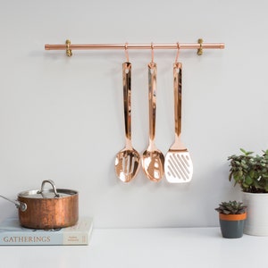 Wall mounted utensil rail made from polished copper, with solid brass fittings and screws. It is holding brass utensils and a pan using the solid copper hooks.