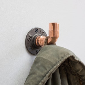 Könnte beinhalten: Ein Haken aus Kupferrohr mit einer schwarzen Metallflansch. Der Haken ist an einer weißen Wand montiert und hat eine Jacke daran hängen.