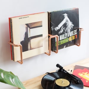 Può includere: Un porta dischi in tubo di rame montato su una parete bianca con due dischi in vinile. Il disco a sinistra è "Dire Straits" e il disco a destra è "Bob Marley and the Wailers -  Easy Skanking - Boston 78".