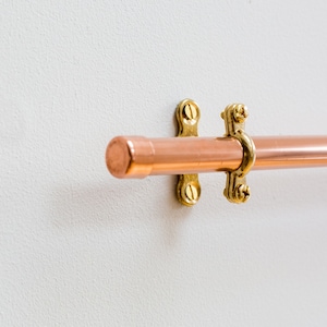 Wall mounted utensil rail made from polished copper, with solid brass fittings and screws. It is holding brass utensils and a pan using the solid copper hooks.