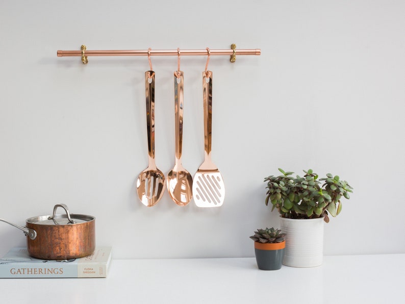 Wall mounted utensil rail made from polished copper, with solid brass fittings and screws. It is holding brass utensils and a pan using the solid copper hooks.