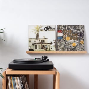 May include: Two vinyl records on a wooden shelf, one with a photo of a house and palm trees and the text "Eric Clapton - 461 Ocean Boulevard", the other with a marbled design and the text "The Stone Roses". A black turntable sits on a wooden table with a stack of vinyl records below.