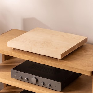 May include: A light wood turntable platform with brass feet sits on a wooden shelf above a black stereo receiver with the word "CAMBRIDGE" on the front.