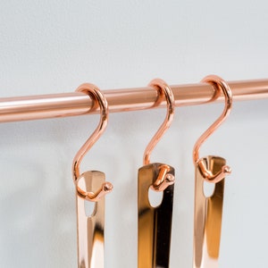 Wall mounted utensil rail made from polished copper, with solid brass fittings and screws. It is holding brass utensils and a pan using the solid copper hooks.
