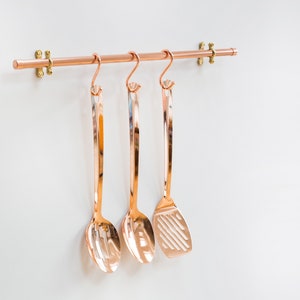 Wall mounted utensil rail made from polished copper, with solid brass fittings and screws. It is holding brass utensils and a pan using the solid copper hooks.