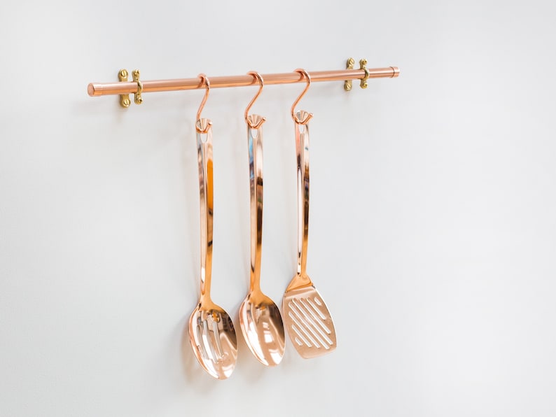 Wall mounted utensil rail made from polished copper, with solid brass fittings and screws. It is holding brass utensils and a pan using the solid copper hooks.