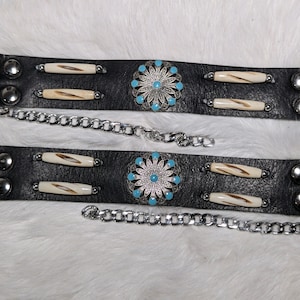 Puede incluir: Dos lazos de cinturón de cuero negro con detalles metálicos plateados y diseños florales turquesa y blancos. Cada lazo tiene una cadena plateada.