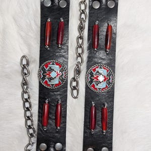 May include: Black leather belt straps with silver-tone hardware and decorative red beads. Each strap features a circular, multi-colored emblem with a Southwestern design. Silver chains connect the straps.