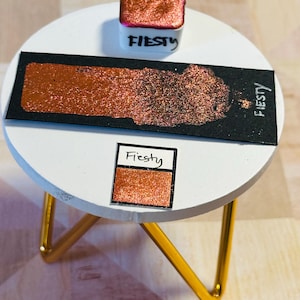May include: A small sample of copper-colored glitter pigment with a black label that says "FIESTY" in white lettering. The sample is on a white round table with a gold metal stand.