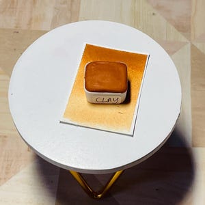 Op de afbeelding: Een klein vierkant bakje met aquarelverf in terracotta kleur. Het bakje is gelabeld met "CLAY" en staat op een wit oppervlak.