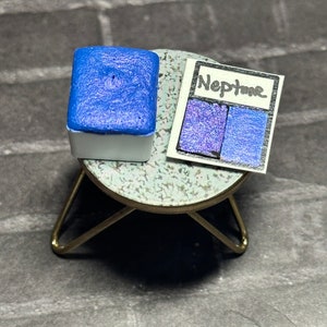 May include: A small, square, blue watercolor paint pan with a white label that says "Neptune" and shows a color swatch of the paint.