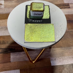 Puede incluir: Un pequeño recipiente cuadrado de pintura iridiscente verde y dorada con la palabra "Jackpot" escrita en la etiqueta. La pintura también se muestra en una superficie cuadrada blanca.