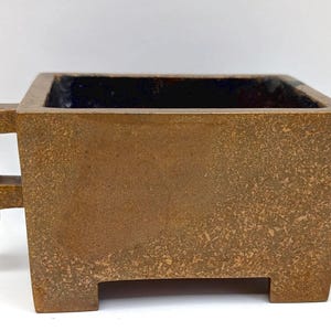 May include: A square, brown, textured container with a dark interior. The container has two rectangular handles on the sides. The design is minimalist and geometric, suitable for decorative purposes.