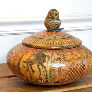 Op de afbeelding: Een decoratieve keramische pot met een deksel, met een uilvormige handgreep. De pot is met de hand beschilderd met illustraties in oude Griekse stijl in tinten oranje, bruin en zwart. De pot staat op een houten oppervlak.