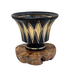 Op de afbeelding: Een zwarte keramische vaas met een gouden geometrisch patroon staat op een houten voet. De vaas heeft een brede opening en een smalle voet.