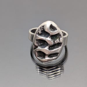 May include: A silver ring featuring a textured oval-shaped design. The design resembles a leaf or a stylized organic shape.
