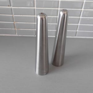 May include: Two stainless steel salt and pepper shakers. The shakers are tall, conical, and have a small hole at the top. They are placed on a gray surface with a tiled background.